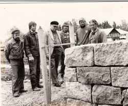 Sex personer, varav mannen i baskern r kyrkoherde Per Olof Persson, betraktar bygget av en stenmur vid kyrkan i Mullhyttan. - klicka fr att frstora