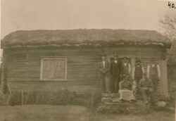 En grupp m�nniskor och en hund p� trappan till en enkel byggnad med torvtak d�r bland annat John Grytsell anges finnas med - klicka f�r att f�rstora