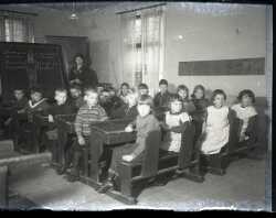 En grupp skolbarn med en lärarinna vid en svart tavla i bakgrunden. Bilden kan vara tagen i småskolan i Hackvads Via - klicka för att förstora