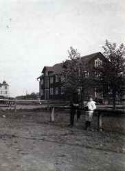 Magasinsbyggnad vid järnvägsstationen med två personer. I bakgrunden till vänster syns hotellet. - klicka för att förstora